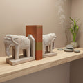 A pair of carved wooden elephant bookends holding a book, displayed on a wooden shelf next to a grey stones and burning incense