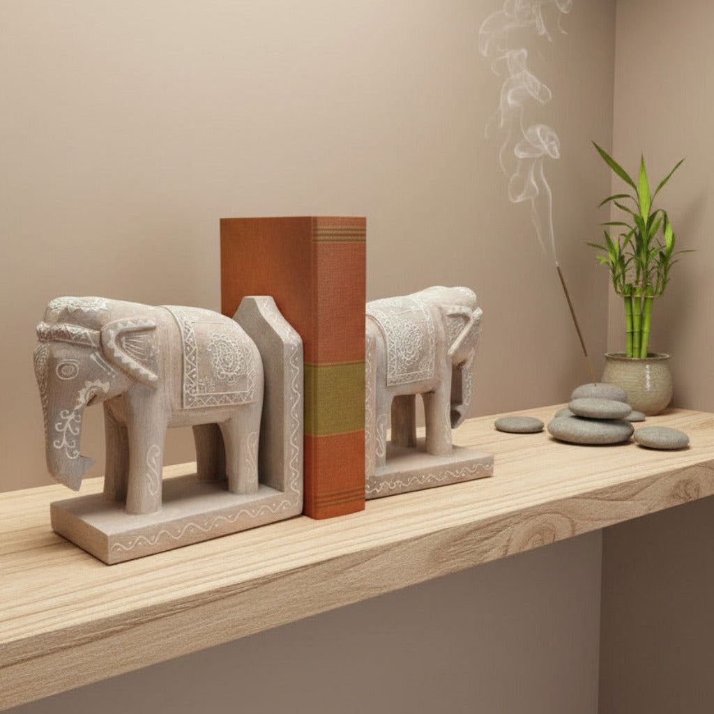 A pair of carved wooden elephant bookends holding a book, displayed on a wooden shelf next to a grey stones and burning incense