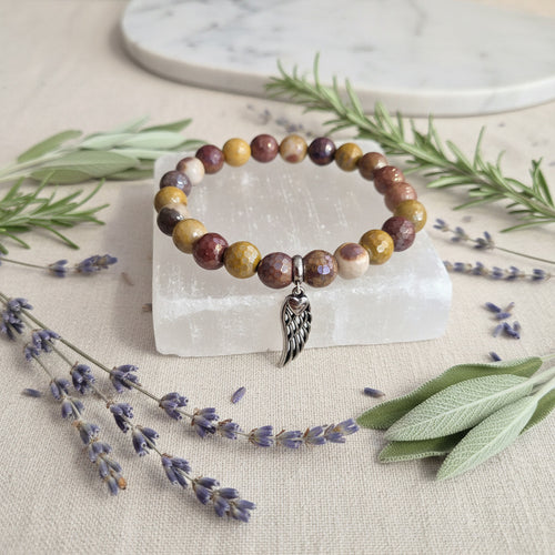 Multicolored beaded bracelet with a silver wing charm on a clear stone block, surrounded by lavender and greenery.