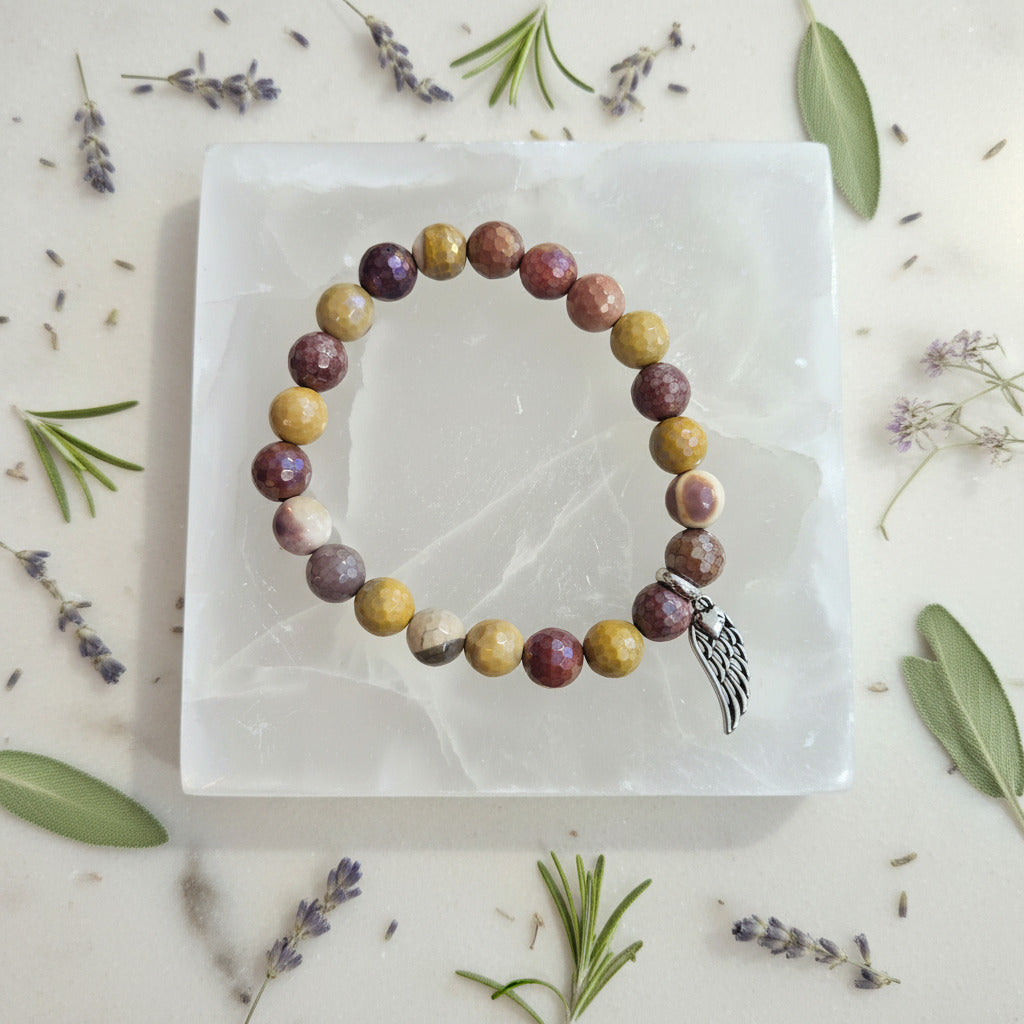 Multicolored beaded bracelet with a silver angel wing charm on a white stone surface, surrounded by herbs.
