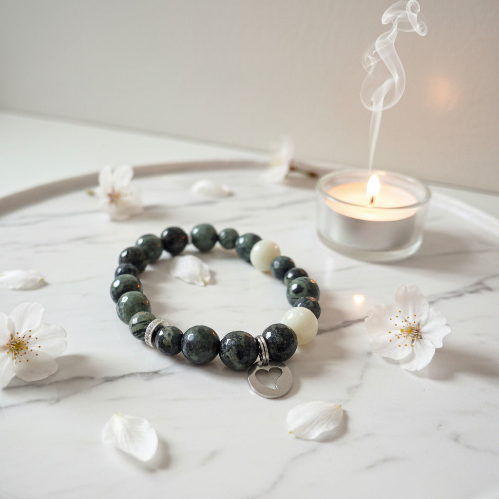 Beaded bracelet with heart charm on a marble surface with flowers and a candle.