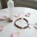 A beaded stretch bracelet made of pink, white, stone beads, with a small silver wing charm, displayed on a marble surface with rose petals,a selenite tower and a tumbled stone.

