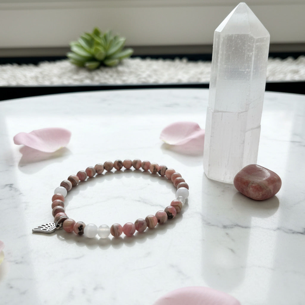 A beaded stretch bracelet featuring pink and white stone beads, and a small silver-toned angel wing charm, displayed on a marble surface.
