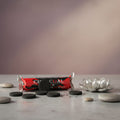 A packaged bar of charcoal hookah fuel wrapped in red, black, and white foil, placed next to a beige incense burner with cone incense burning on a light beige surface with stones around