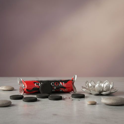 A packaged bar of charcoal hookah fuel wrapped in red, black, and white foil, placed next to a beige incense burner with cone incense burning on a light beige surface with stones around