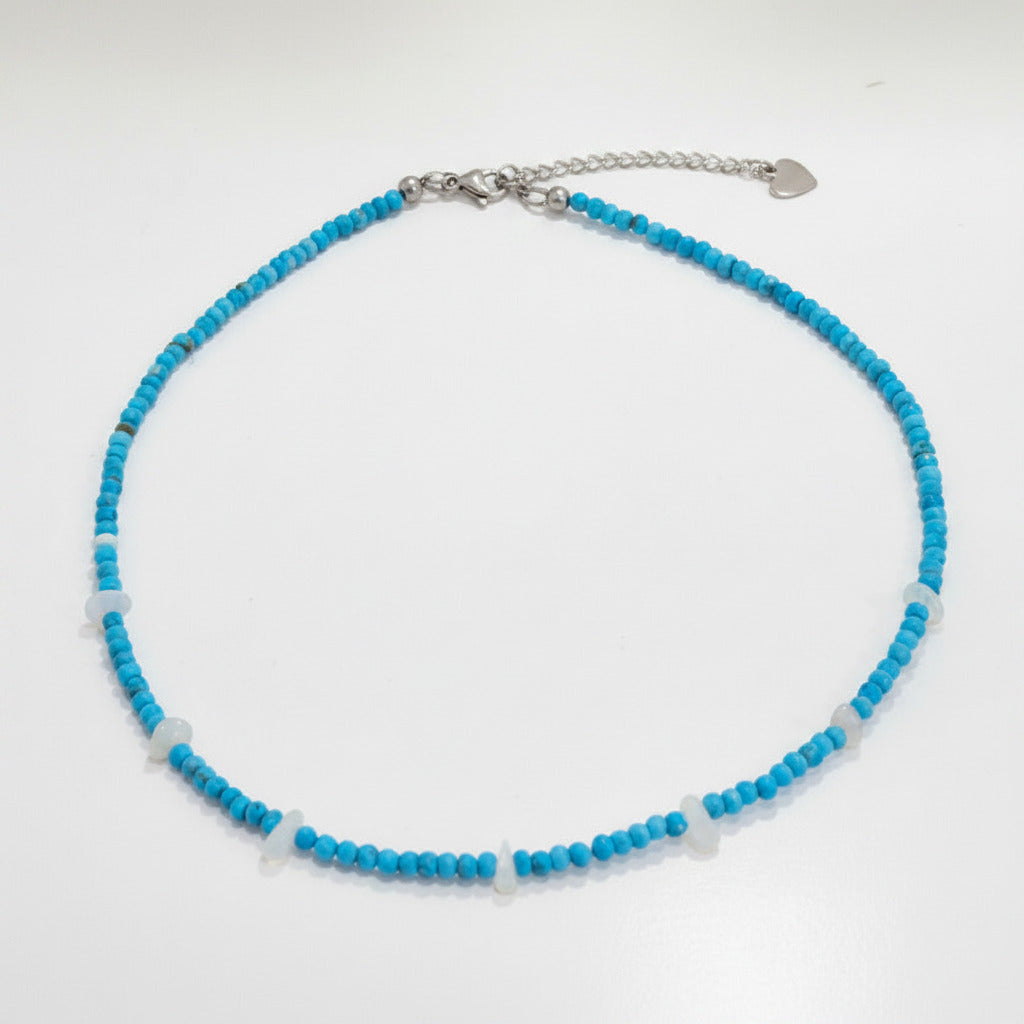 A beaded necklace with small turquoise-blue beads, accented by a few translucent white beads and a silver-tone clasp with an extender chain and small heart charm, laid flat on a light surface.
