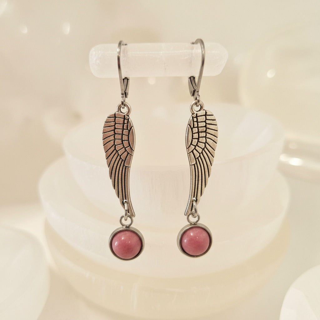 Silver color wing-shaped earrings with pink stones on a white stand.