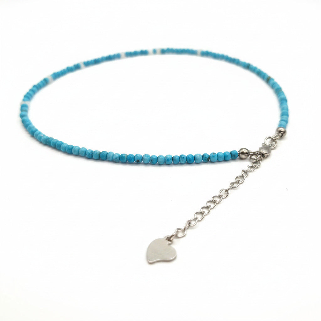 A beaded necklace with light blue round beads, a silver-tone chain extender, and a small silver heart-shaped charm, laid on a white fabric background.