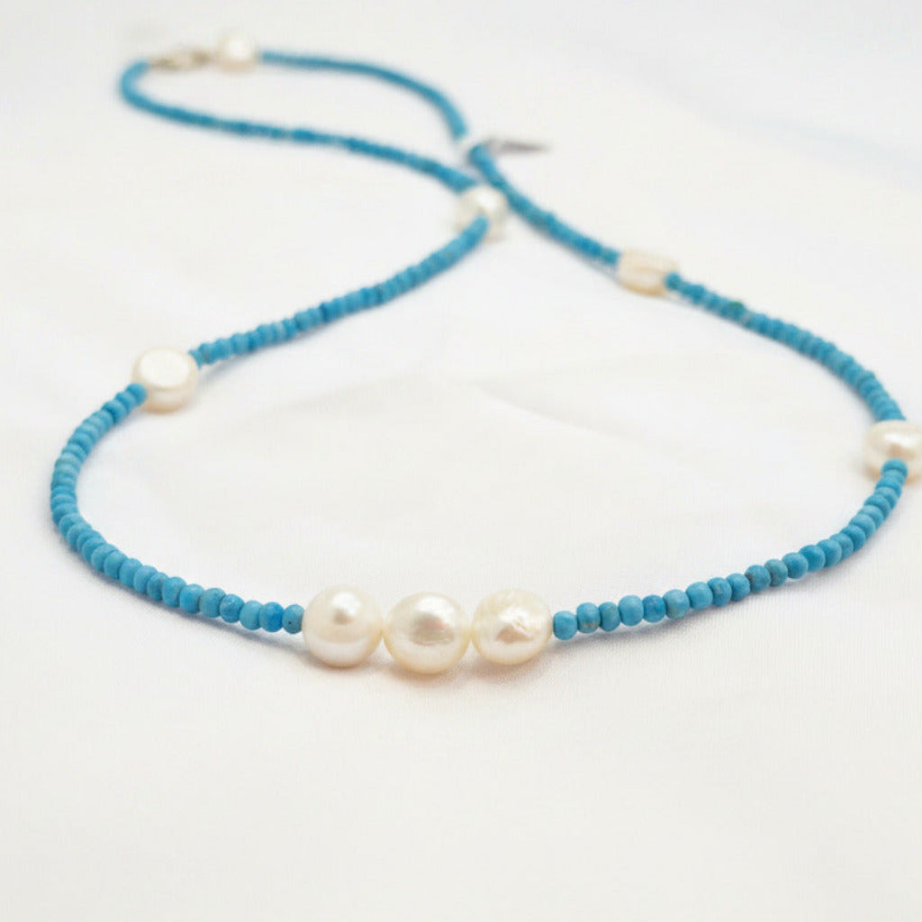 A beaded necklace with bright turquoise-blue beads and irregular white freshwater pearls, displayed on a white fabric surface.