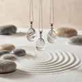 Three silver necklaces with teardrop-shaped quartz pendants on a sandy background with stones.