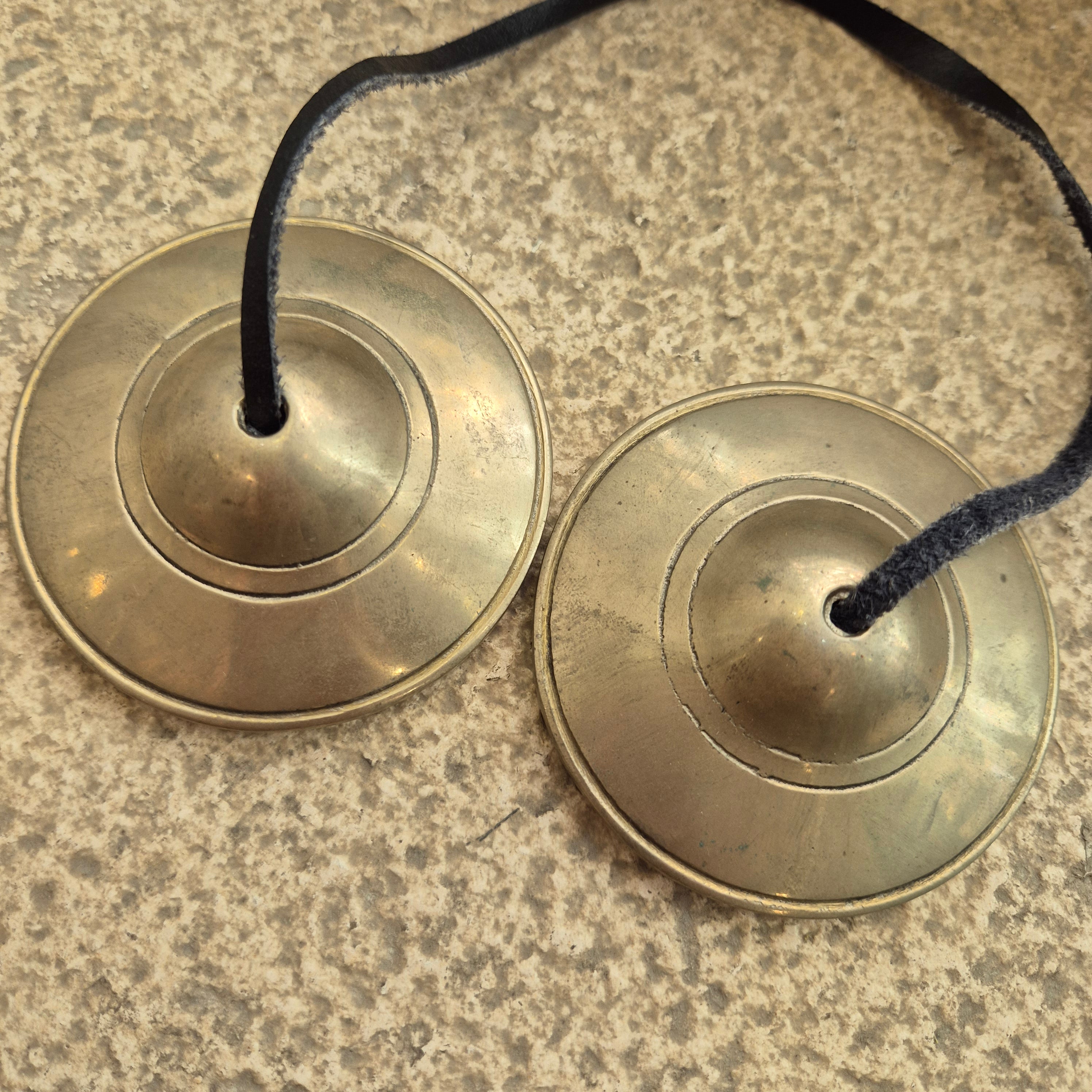 A pair of small brass-toned cymbals or hand clackers with black cords, photographed on a speckled stone-like surface.