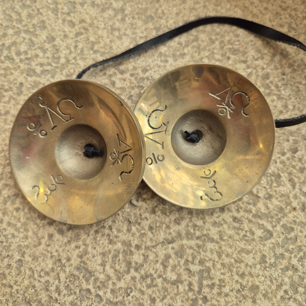 A pair of small brass cymbals with engraved symbols, connected by a black cord, resting on a textured stone surface.