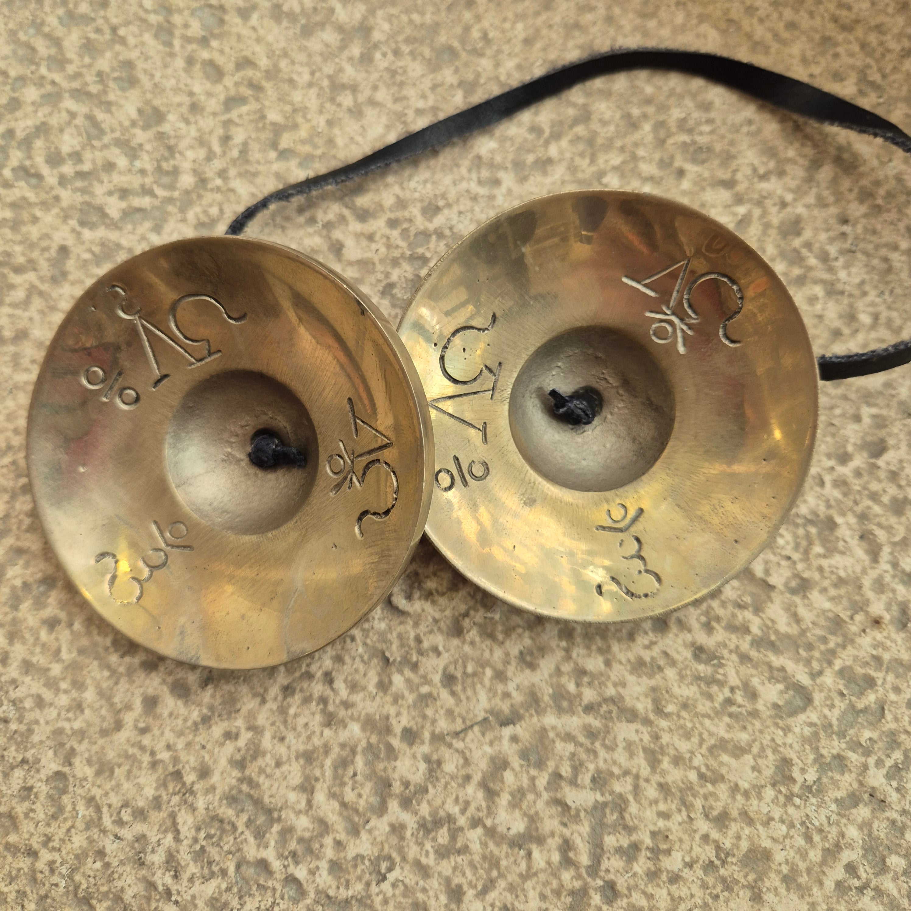 A pair of small brass cymbals with engraved symbols, connected by a black cord, resting on a textured stone surface.