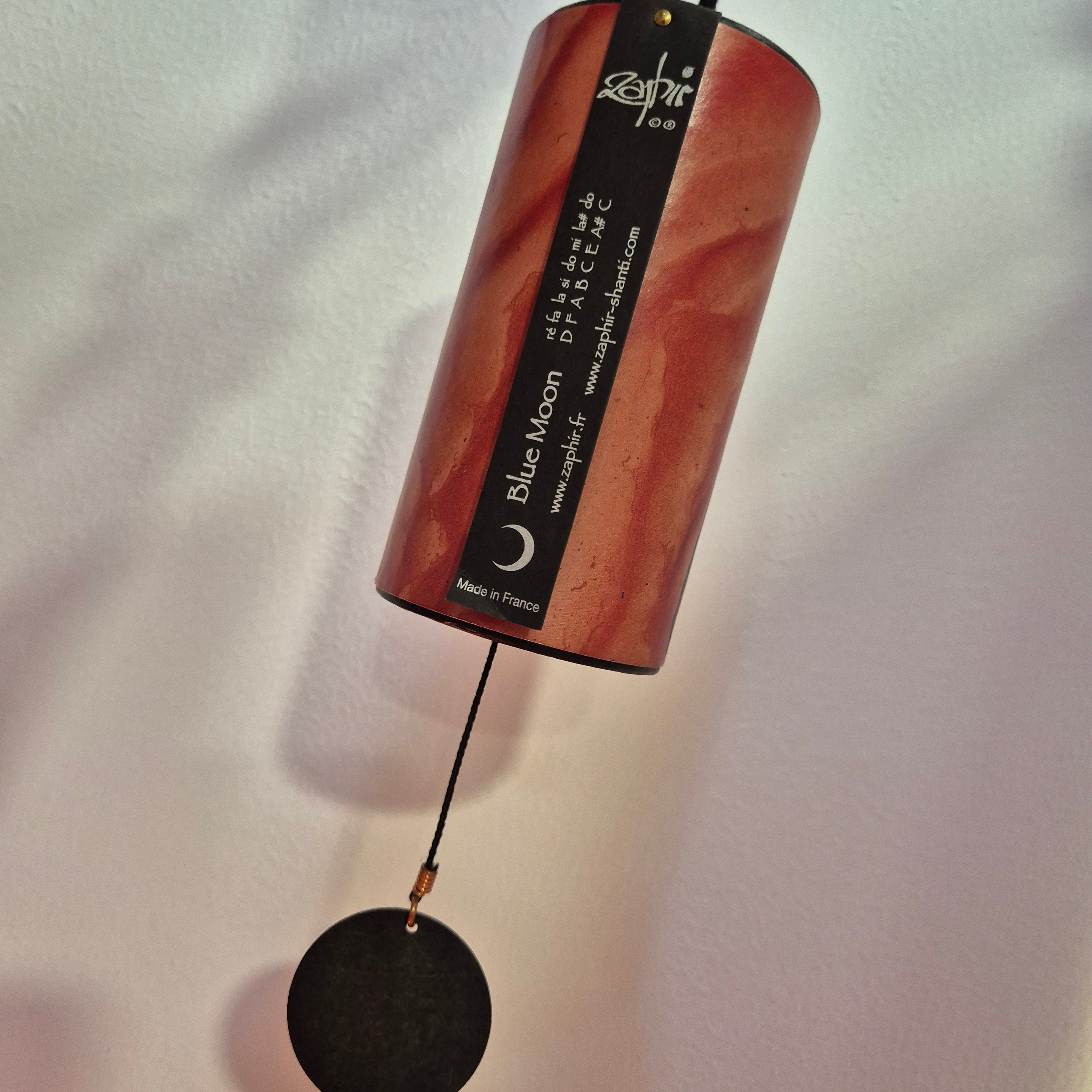 A cylindrical wind chime bell with a marbled reddish-brown and orange finish, hanging from a black cord with a round black striker. The bell features a black label with white text reading 'blue moon' and 'appliance'.