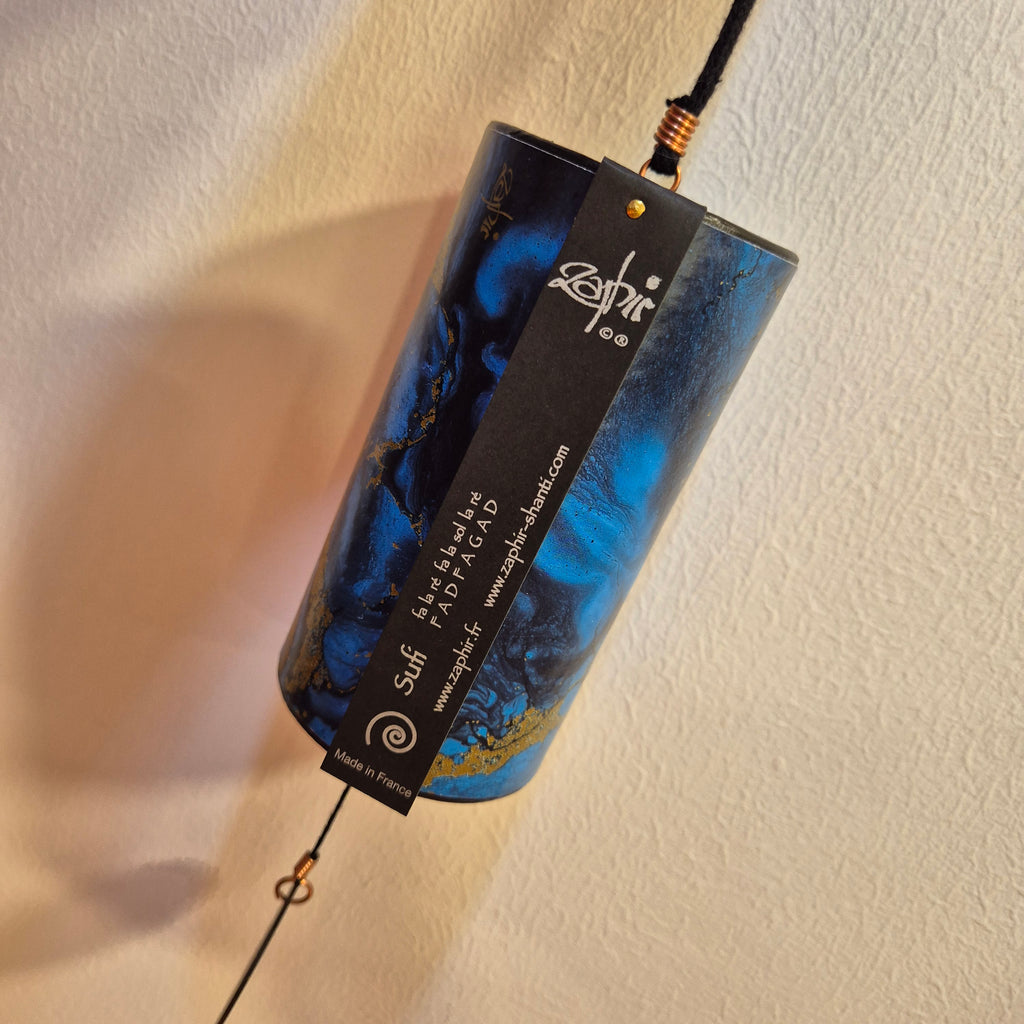A cylindrical wind chime blue and black marbled pattern and gold accents, hanging against a textured beige wall. A black hanging tag with the brand name 'Suff' is attached to the front