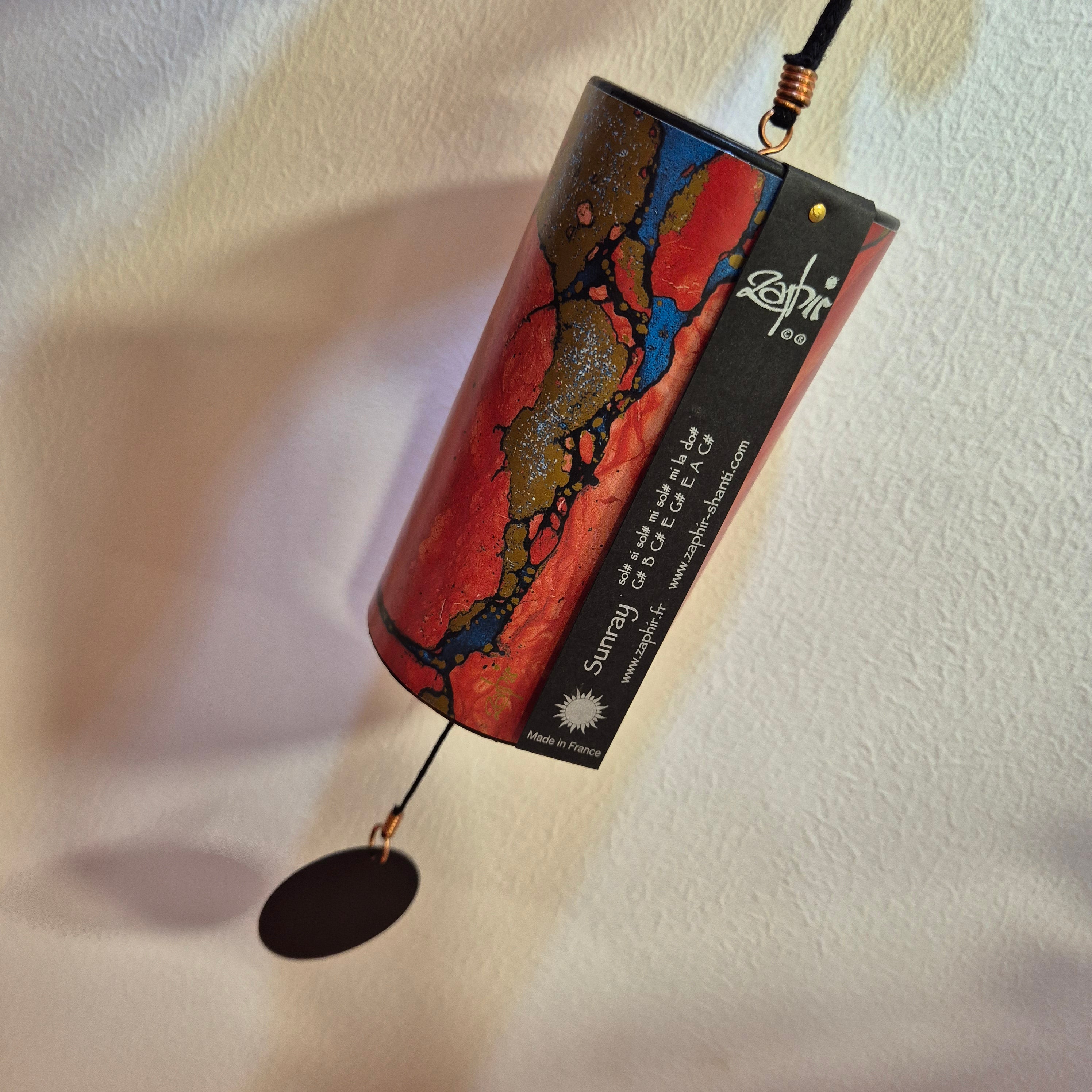 A cylindrical hanging wind chime with a marbled red, orange, black, and blue design, featuring a black side label with white text and a dark oval striker at the bottom, photographed against a textured off-white wall.