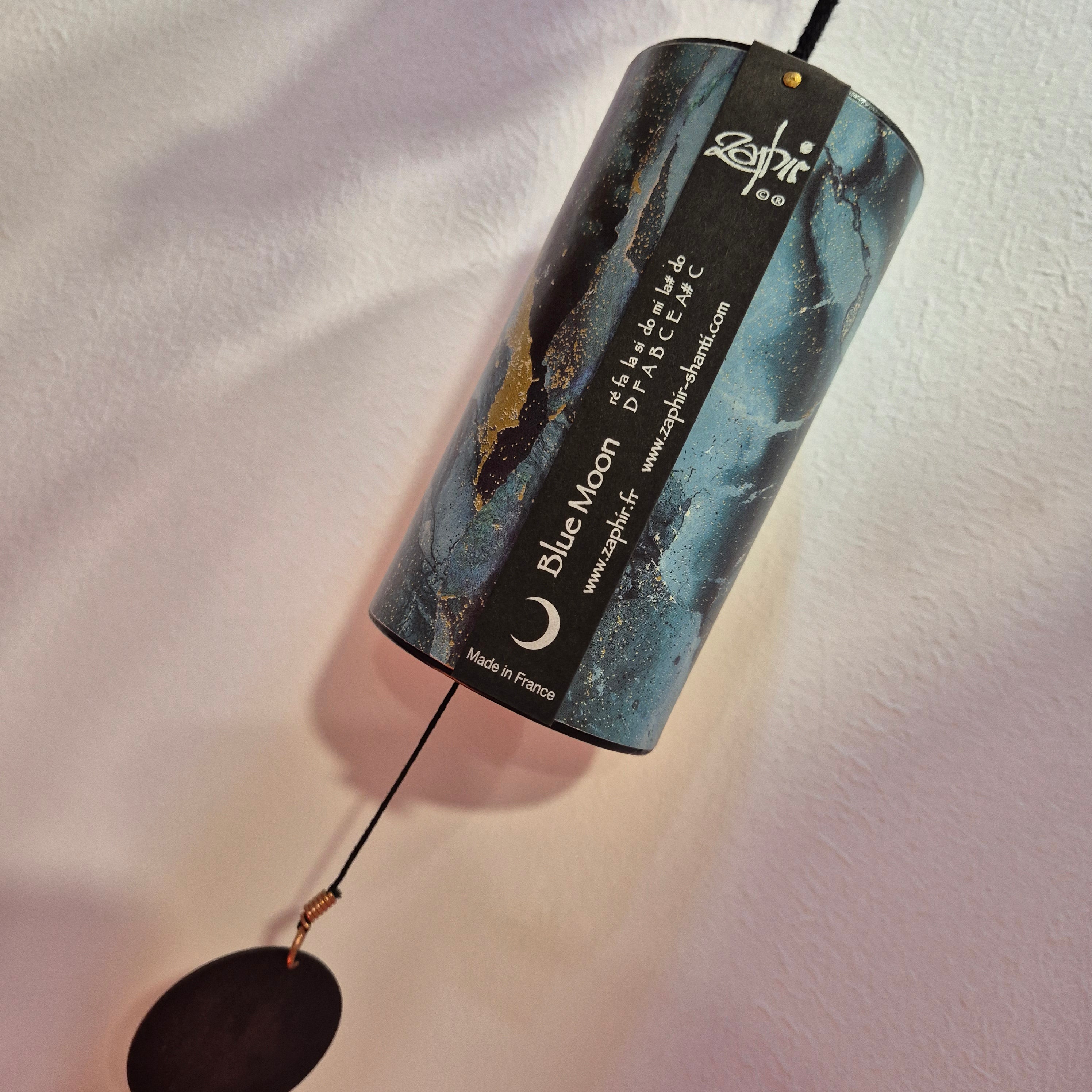 A cylindrical blue and black marble wind chime labeled 'Blue Moon' by Zenith, hanging against a textured white wall with a dark metal striker and cord below.
