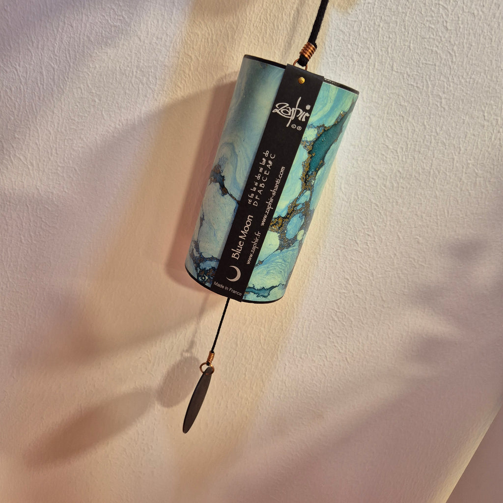 A cylindrical Zaphir Blue Moon wind chime with a blue and green marbled pattern, hanging from a black cord against a textured beige wall.