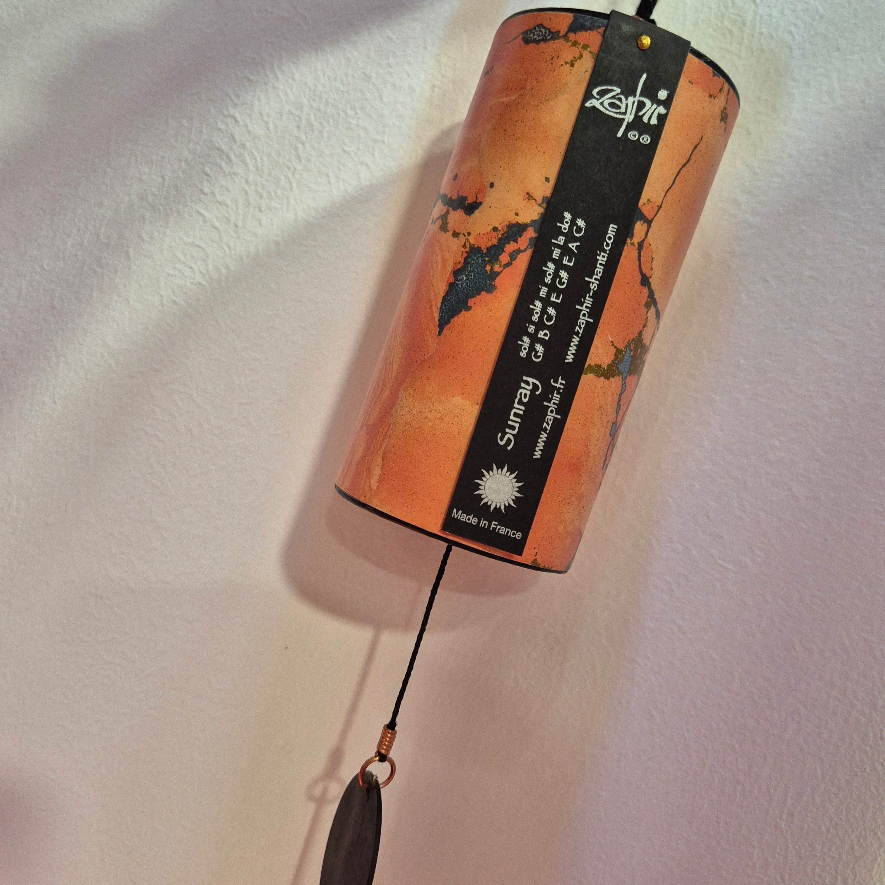 A cylindrical wind chime hanging from a black cord against a textured off-white wall. The chime has an orange and black marbled pattern, with a black vertical label that reads 'Zenith' and 'Sunray Wind Chime'.
