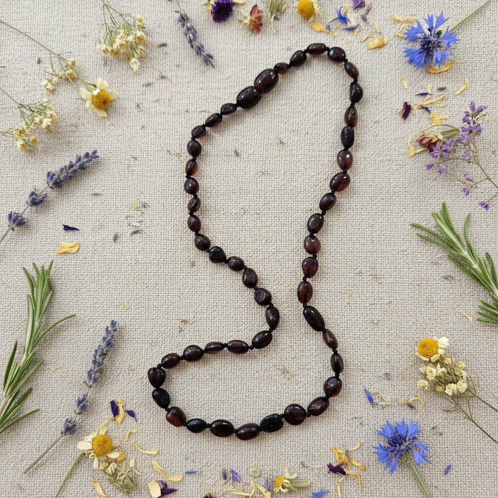 A beaded necklace made of dark, polished oval amber beads  on a beige surface surrounded by dried herbs