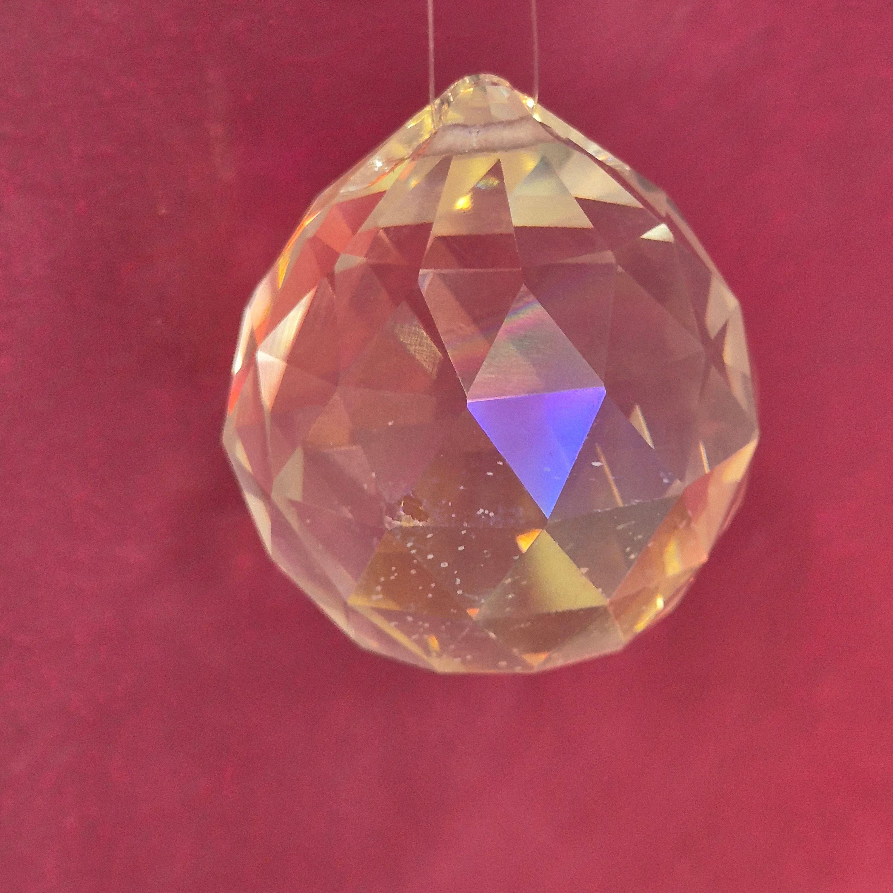 A faceted round-shaped clear glass crystal bead with an iridescent finish, resting on a textured dark red background.