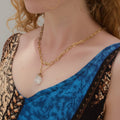 Person wearing a gold necklace with a crystal pendant against a pink background