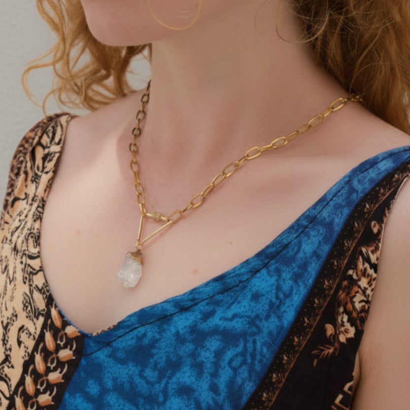 Person wearing a gold necklace with a crystal pendant against a pink background