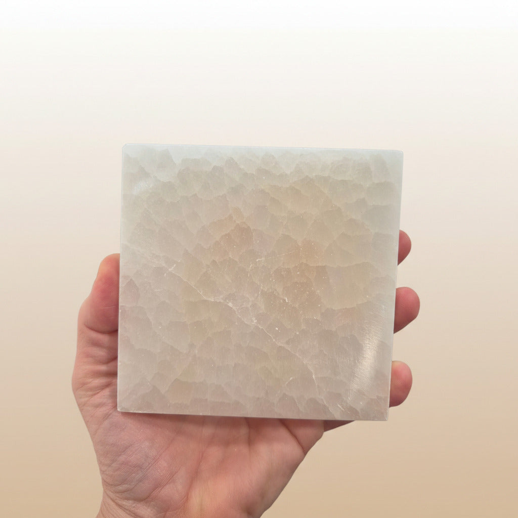 Hand holding a square selenite plate with a beige marble pattern against a plain background