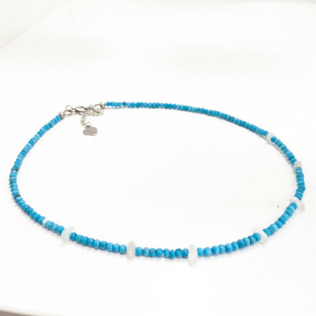 A beaded necklace featuring bright turquoise blue beads and a few translucent white beads, with a silver-tone clasp and heart charm, displayed on a white surface.