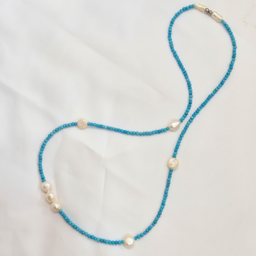 A beaded necklace featuring bright turquoise blue beads and irregular white pearl-like accents, laid out on a white fabric background.