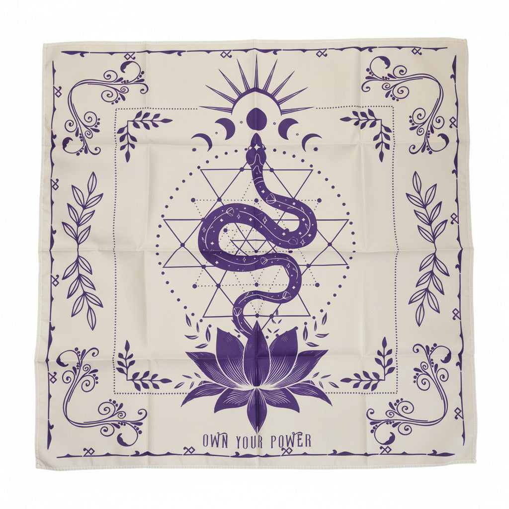 A square cream-colored cloth or scarf laid flat on a light wood floor, featuring a purple occult-style design with a snake, lotus flower, geometric symbols, and the text 'OWN YOUR POWER'.